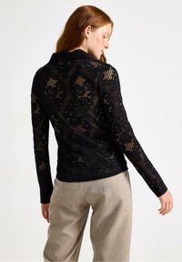 Svart spetsblus med långa ärmar och blommönster, med krage och en något figursydd form. Bärs över ljusbeige byxor.