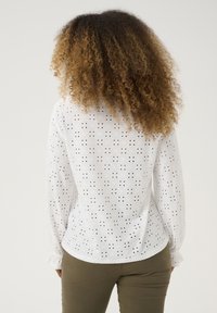 Camisa blanca de manga larga con intrincados patrones recortados, con un dobladillo redondeado y puños fruncidos, combinada con pantalones verde oliva.