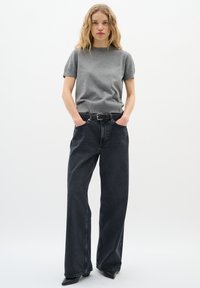 Junge Frau mit welligem blonden Haar, die ein graues Kurzarmoberteil, schwarze weite Jeans, einen schwarzen Gürtel und schwarze spitze Schuhe trägt, steht mit den Händen in den Taschen.