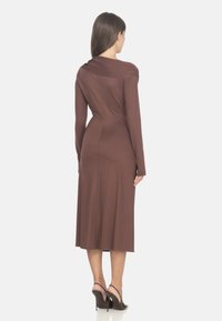 Abito lungo marrone a maniche lunghe con scollatura drappeggiata e silhouette aderente, che arriva a metà polpaccio. Il tessuto è liscio con una leggera lucentezza.