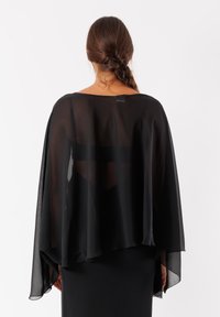 Overlay in tessuto nero trasparente a forma di mantello con drappeggio fluido, design aperto e bordi morbidi, si adagia su un indumento aderente, rivelando una silhouette semplice.