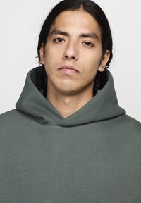 Jovem com cabelo escuro de comprimento médio, vestindo um sweatshirt cinza com capuz, olhando diretamente para a câmara com uma expressão neutra.