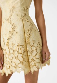 Robe jaune sans manches avec des découpes en dentelle florale complexe et un ourlet festonné, portée par une personne les mains détendues le long du corps.