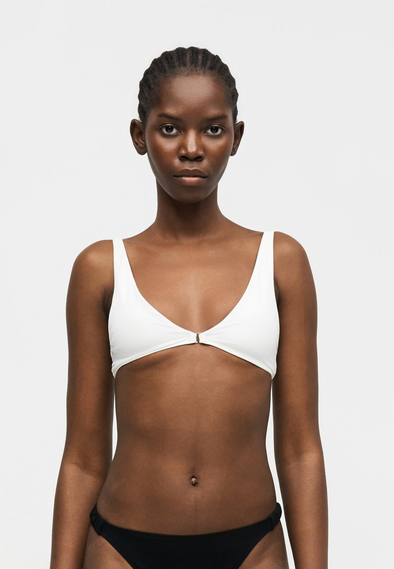 Γυναίκα με πλεξούδες, φορούν λευκό μπικίνι με κούμπωμα μπροστά και μαύρο κάτω μέρος μπικίνι, στέκεται μπροστά σε απλό λευκό φόντο.