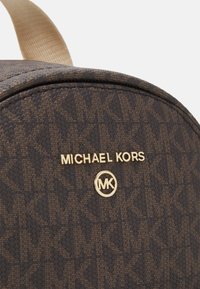 Brauner Rucksack mit Muster, gefertigt aus strukturiertem Material, mit goldfarbener Markierung und einem kreisförmigen Logo. Akzentriemen in heller Farbe.
