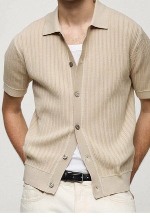 Beige kortærmet strikket skjorte med knapper, båret åben over en hvid tanktop, kombineret med hvide bukser og sort bælte.