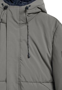 Chaqueta acolchada gris con un alto cuello, capucha con cordón y bolsillos con cremallera. Presenta un tejido suave y un forro interior acolchado.