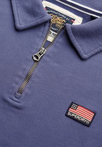 Sudadera azul con cremallera de cuarto de vuelta y cremallera de metal, con un parche bordado de la bandera americana etiquetado "SUPERDRY" en el pecho.