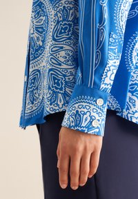 Camicia blu con motivo a paisley bianco, maniche lunghe con polsini a bottone e una texture di stoffa liscia.