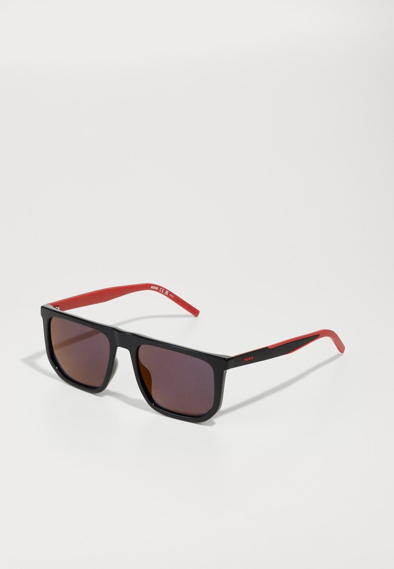 Ochelari de soare pătrați negri cu un finisaj lucios, brațe roșii și lentile maro cu gradient. Au un logo pe brațul lateral și un design elegant.
