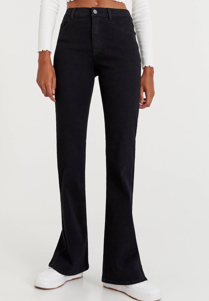 Jean évasé noir en denim, avec une taille haute et un design à cinq poches. Associé à des baskets blanches. Texture propre et minimaliste.