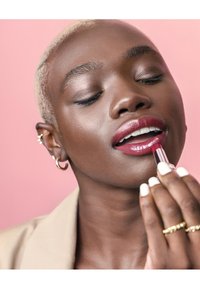 Rouge à lèvres dans un tube métallique appliqué sur les lèvres. Le modèle a les cheveux blonds courts, porte des boucles d'oreilles créoles et des clous, avec une manucure neutre et des bagues.
