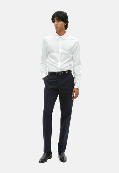 Mężczyzna stoi z rękami w kieszeniach, ubrany w białą koszulę, czarny pasek, granatowe spodnie i czarne buty wizytowe na jednolitym tle.