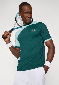 Camisa de tenis en verde y azul claro con cuello redondo, mangas cortas y logo de EA7. Fabricada con material transpirable, presenta un diseño deportivo.