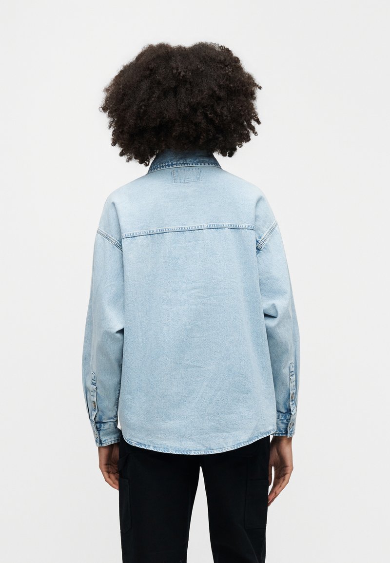 Light blue denim jacket with a relaxed fit, button cuffs, and a curved hem. Features a back yoke and no visible embellishments.