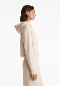 Donna in felpa a maglia beige e gonna lunga abbinata, rivolta a destra, su uno sfondo bianco.