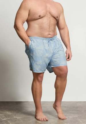 Mężczyzna bez koszulki w jasnoniebieskich kąpielówkach z motywem smoków, stojący boso na teksturowanej szarej podłodze na jednolitym tle.