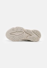 Vaalean beige kenkäsole, jossa on teksturoitu kumipinta, kaarevat viivat ja pyöreät yksityiskohdat. Sisältää "adidas"-merkin lähellä kantapäätä.