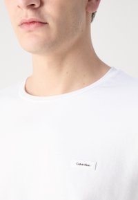 Hvit bomulls t-skjorte med rund hals, med en liten rektangulær Calvin Klein-etikett på venstre bryst.