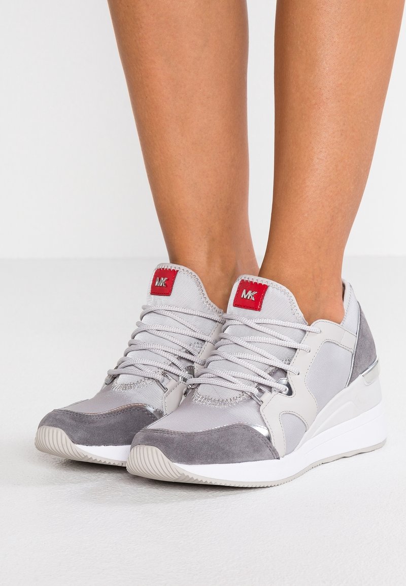 Baskets compensées gris clair et argent avec embouts en daim et écussons rouges avec logo sur les languettes, portées pieds nus sur un fond blanc.