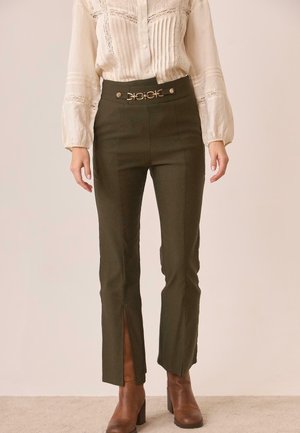 Mujer con pantalones de talle alto en verde oliva con aberturas delanteras y cinturón decorativo con hebilla dorada, combinados con una blusa de encaje color crema y botas marrones.