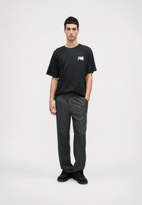 Μαύρη oversized μπλούζα με γραφικό σχέδιο, γκρι παντελόνι με ρίγες και μαύρα αθλητικά παπούτσια. Μοντέλο στέκεται σε ουδέτερο φόντο.