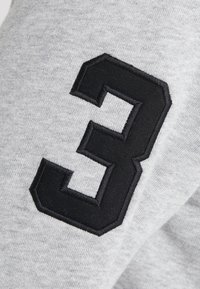 Una tela gris con una textura suave presenta un gran número "3" bordado en negro en la manga, contorneado por un borde texturizado.