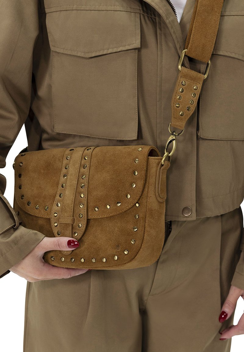 Bruine suède crossbody tas met metallic studs, rechthoekige vorm, gevouwen klep en verstelbare schouderband. Draagt boven een kaki outfit.