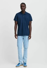 Mornarska polo majica s kratkimi rokavi, ovratnikom in diskretnim logotipom, v kombinaciji s svetlo modrimi jeans hlačami in modrimi supergami z belimi črtami.