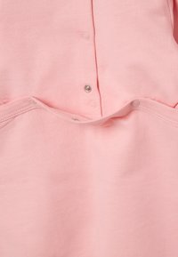 Rosa Baumwollkleidung mit Druckknöpfen an der Vorderseite und einer weichen, glatten Textur. Einschließlich Taschen mit gebogenen Öffnungen.