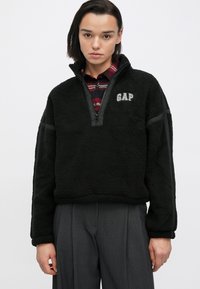Pullover in pile nero con collo con zip, presenta un piccolo logo GAP sul petto e dettagli neri a contrasto lungo le maniche.