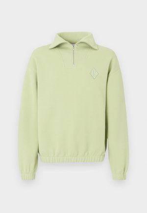 Sweatshirt vert clair avec un col zippé à mi-hauteur, texture côtelée, taille élastique et un logo en forme de diamant sur la poitrine.