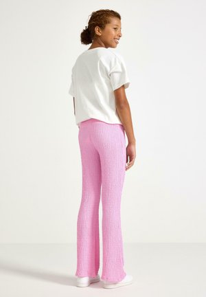 Dívka se usmívá, má na sobě bílé tričko s krátkými rukávy a růžové texturované kalhoty, stojí před jednoduchým světlým pozadím.