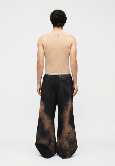 Άνδρας με μαύρα σγουρά μαλλιά φοράει μπεζ αμάνικη μπλούζα και φαρδιά μαύρη παντελόνα με καφέ batik σχέδια, στέκεται γυρισμένος πλάτη σε λευκό φόντο.