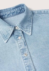 Chemise en denim bleu clair avec col pointu et trois boutons-pression argentés visibles sur la bande de boutonnage.
