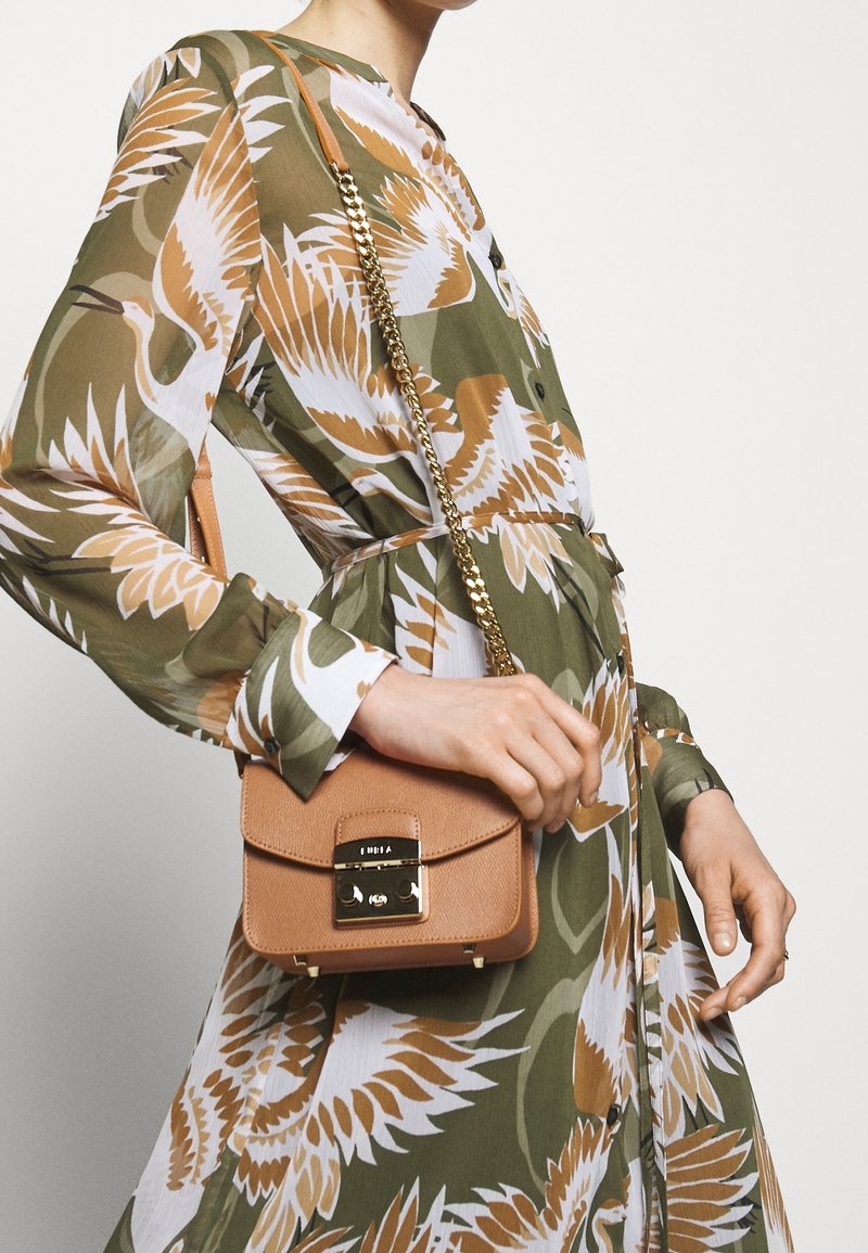 Femme portant une robe florale verte et beige tenant un petit sac en cuir marron clair à bandoulière avec chaîne et fermoir dorés.