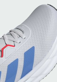 Baskets de sport gris clair avec tige en maille texturée, présentant des rayures bleues et des accents rouges, bout arrondi et système de laçage.