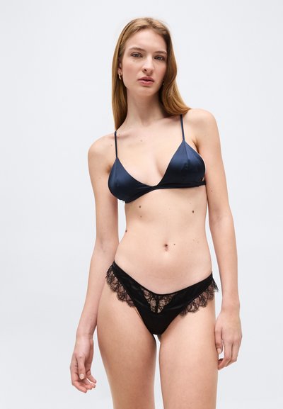 Γυναίκα που φοράει μπλε σατέν σουτιέν με λεπτές τιράντες και σατέν εσώρουχο με μαύρη δαντέλα, στέκεται μπροστά σε ένα απλό ανοιχτόχρωμο φόντο.