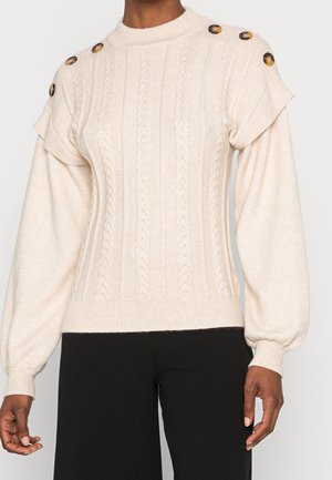 Pull crème en maille torsadée avec boutons aux épaules et manches longues bouffantes, porté avec un pantalon noir.