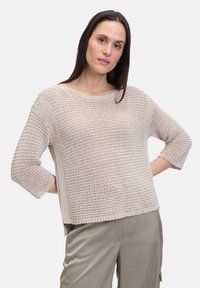 Beige gebreide trui met een los vallende pasvorm, driekwartmouwen, ronde halslijn en een textuurpatroon. Gecombineerd met lichte, neutrale broeken.