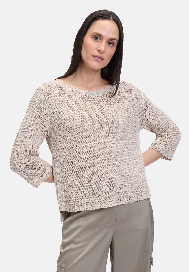 Beige gebreide trui met een los vallende pasvorm, driekwartmouwen, ronde halslijn en een textuurpatroon. Gecombineerd met lichte, neutrale broeken.