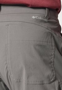 Pantalon d'extérieur gris Columbia avec coutures sur la poche arrière et logo, associé à une chemise rouge rentrée à la taille.