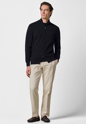 Hombre de pie con las manos en los bolsillos, vestido con un suéter negro con cremallera, pantalones beige y mocasines marrón oscuro, frente a un fondo liso.