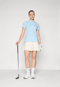 Šviesiai mėlyna polo marškinėliai su apykakle, trumpomis rankovėmis ir baltais akcentais, derinami su kremine klostuota suknele. Matomos baltos bateliai ir golfų klubas.