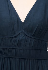 Κοντινό πλάνο ενός σκουρομπλε φορέματος με ντεκολτέ σε σχήμα V, ρεταρισμένο ύφασμα και λεπτομέρεια με ελαστική μέση.