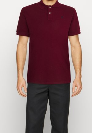 Mężczyzna w ciemnoczerwonej koszulce polo z krótkim rękawem, z dwoma guzikami, oraz czarnych spodniach, stojący na jednolitym tle.