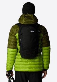 Svart ryggsekk med polstret design brukt over en grønn pufferjakke, med rund topp, logo detaljer og glatt tekstur.