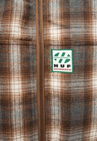 Veste à carreaux marron et grise avec une fermeture éclair marron et un patch blanc bordé de vert lisant "HUF San Francisco 02" sur la poitrine.