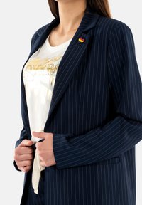 Blazer a righe navy con revers a notched, due tasche frontali e una t-shirt bianca con grafica e dettagli metallici. Piccolo distintivo attaccato.