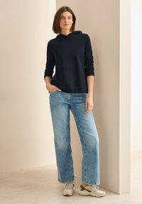 Sudadera azul marino, material suave, corte holgado, mangas largas. Jeans de pierna ancha azul claro, aspecto desgastado, combinados con zapatillas blancas y grises.